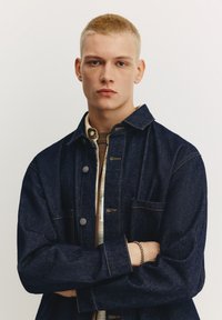 Jeans jakna temno modre barve z vidnimi šivi, kovinskimi gumbi in žepom na prsih. Nosi se prek lahke karo srajce z rjavim pletenim ovratnikom.