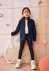 Giacca in pile nera con zip, maglietta bianca, leggings neri, scarpe da ginnastica beige, che tiene in mano una racchetta da tennis bianca contro una parete in legno chiaro.