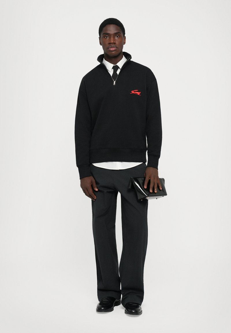 Man in een zwarte sweatshirt met kwartrits, witte shirt, zwarte stropdas, donkere broek, met een zwarte glanzende clutch in de hand, staande tegen een witte achtergrond.