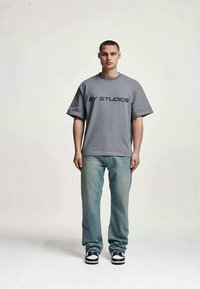Um jovem homem está de pé, virado para a frente, vestindo uma t-shirt cinza da "2Y Studios", calças de ganga azul claro e ténis preto e branco, contra um fundo simples.