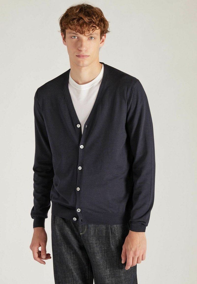 Cardigan marine en tissu doux, doté de manches longues, d'un décolleté en V et de six boutons blancs sur le devant. Porté par-dessus une chemise blanche.