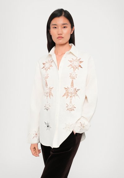 White button-up shirt featuring intricate star patterns in cut-out lace. Paired with dark pants. Smooth texture and relaxed fit.