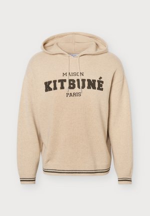 Мъж позира с бежов суичър с качулка Maison Kitsuné Paris, широки сини дънки и кафяви обувки на изчистен светъл фон.