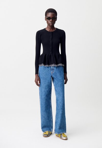 Persona alta che indossa una camicia nera a maniche lunghe con bottoni e orlo arricciato, jeans blu a gamba larga, scarpe da ginnastica argentate con lacci gialli e occhiali da sole scuri.