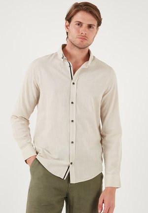 Beige linned skjorte med lange ærmer, knaplukning foran, kontrasterende krave detaljer og subtil tekstur. Sammen med grønne bukser.