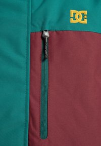 Sección de chaqueta en verde azulado y burdeos con una costura diagonal, que presenta un bolsillo con cremallera y un logo amarillo de DC en la esquina superior.