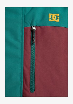 Giacca teal e bordeaux con una cucitura diagonale, dotata di una tasca con zip e un logo DC giallo nell'angolo superiore.