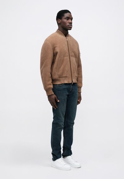 Férfi bézs színű, cipzáros bomberdzsekit visel, bordázott mandzsettákkal, sötétkék farmernadrággal és fehér sportcipővel, egy sima fehér háttér előtt áll.