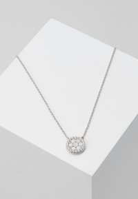 Silver halsband med cirkulär medaljong, prydd med glittrande stenar, på en fin kedja. Vit och metallisk färgpalett.