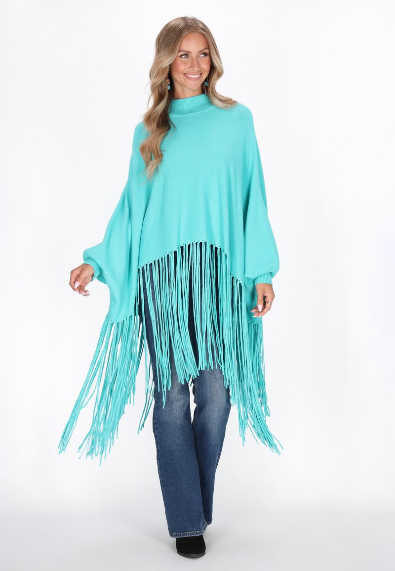 Türkiissinine poncho pikemate äärtega, millel on kõrge kaelus, mugavad varrukad ja lõtv lõige, millele on kombineeritud sinised teksad ja mustad jalanõud.