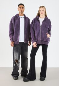 Felpe viola con zip e tasche frontali, indossate sopra camicie bianche. Jeans larghi neri con effetto vissuto e pantaloni a campana neri aderenti. Scarpe nere chunky.