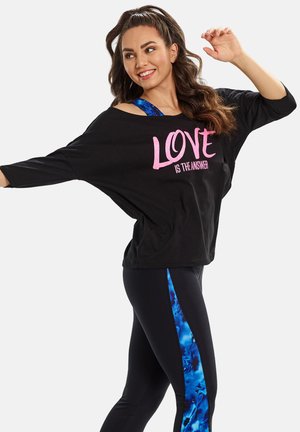 Schwarzes Oversized-Shirt mit pinkem Text "LIEBE IST DIE ANTWORT", kombiniert mit schwarzen Leggings, die an den Seiten ein blaues abstraktes Muster aufweisen.