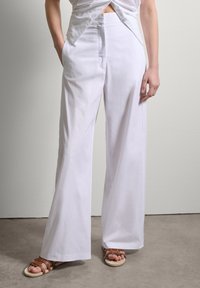 Weiße weite Hosen aus Baumwolle mit glatter Textur, ausgestattet mit Seitentaschen und einer hohen Taille. Kombiniert mit braunen Riemchensandalen.