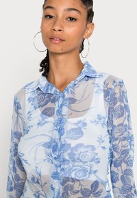 Blusa azul clara transparente com padrão floral, com botão frontal, gola e mangas longas. Apresenta um tecido texturizado com um brilho subtil.