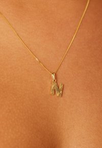 Gouden hangertje met de letter 'M' in reliëf, hangend aan een fijne gouden ketting, tegen een gebruinde huid als achtergrond.