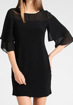 Femme portant une robe noire longueur genou avec manches cloche transparentes superposées et panneau transparent à l'encolure, sur fond uni.