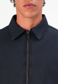 Navy zip-up jakke med krage, med en metallikk glidelås og teksturert stoff. Sømdetaljer langs kragen og kanten.