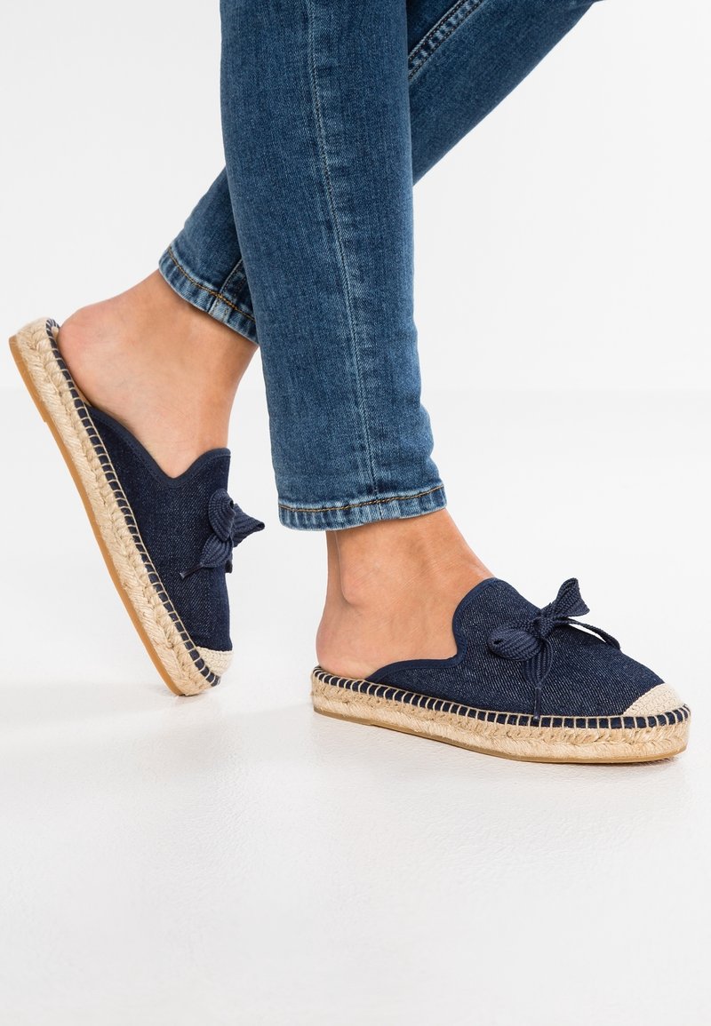 Mules espadrilles navy avec un design à bords festonnés et un accent en nœud, dotés d'une semelle en jute et d'un dessous en caoutchouc. Tige en textile bleu foncé.