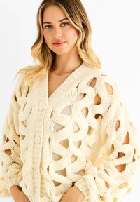 Knit cardigan in cream, featuring an open weave design, long sleeves, and button closure. Textured fabric with wave patterns and ribbed cuffs.