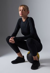 Crni sportski outfit s dugim rukavima i tajicama, sjedi na crnoj i žutoj medicinskoj lopti, s crnim tenisicama. Glatka tekstura.