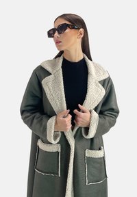 Übergrößiger grüner Mantel mit einem weißen Fleece-Kragen und Taschen, der eine weiche Textur und eine lockere Passform aufweist, getragen über einem schwarzen Oberteil.