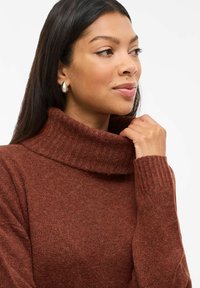 Rjav, teksturiran pulover s turtleneck ovratnikom in rebrastimi manšetami, ki se ponaša z sproščenim krojem. Model nežno drži ovratnik z eno roko.
