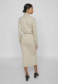 Vestido bodycon de punto beige con cuello alto, mangas largas y silueta ajustada, combinado con zapatos de tacón bloque negro.