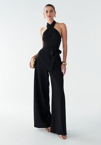 Salopette noire à jambes larges avec encolure halter, tissu texturé et ceinture détachable. Accessoirisée avec une pochette dorée et des bijoux.