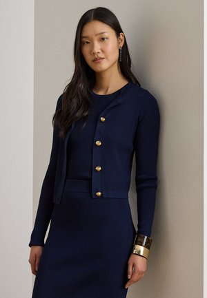 Frau mit langem dunklem Haar, trägt ein marineblaues, geripptes Kleid und eine passende Strickjacke mit goldenen Knöpfen, steht vor einer beigen Wand.