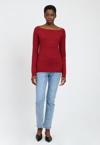 Maglione rosso a maniche lunghe con scollo a barca, design aderente e orlo a coste, abbinato a jeans a gamba dritta azzurri e tacchi neri.