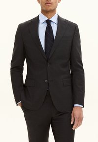 Donkergrijs maatpak met notch-lapels, twee knopen en voorzakken. Draagt een lichtblauw overhemd en een donker gestreepte stropdas.