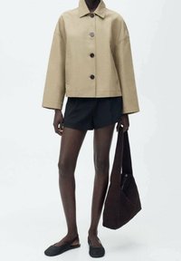 Beige cropped jasje met een kraag en vier zwarte knopen, gecombineerd met zwarte shorts en zwarte gevlochten instappers. Een donkerbruine schoudertas maakt de look af.