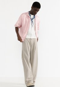 Cămașă roz cu mânecă scurtă, cu un model spiralat, tricou alb, pantaloni bej largi și un fular albastru, purtați cu încălțăminte de culoare deschisă.