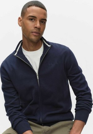 Mörkblå zip-up-jacka med hög krage, vita detaljer, ribbade muddarna och en diskret logotyp på bröstet. Bärs över en vit skjorta.