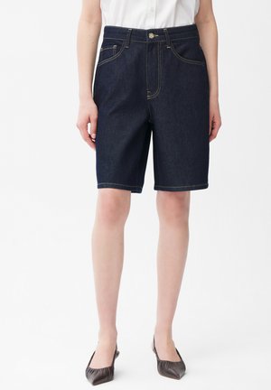 Kvinde iført mørkeblå denim knælange shorts med gul syning, kombineret med hvid skjorte og mørke spidse flade sko.