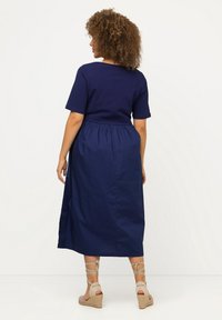 Mörkblå klänning med kort ärm, figurnära överdel och elastisk midja, som flödar ut i en knälång kjol. Buren med kilklackssandaler.