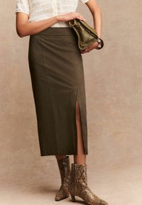 Jupe crayon vert olive avec une fente sur le côté, associée à un chemisier texturé blanc et des bottes en imprimé serpent pour un look superposé. Accentuée par un sac vert matelassé.