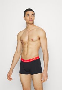 Black boxer shorts with a red elastic waistband featuring "BOSS" logo. Smooth texture, fitted design, mid-thigh length.
