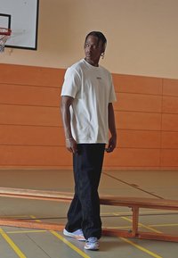 Hvit t-skjorte med korte ermer og en liten logo, sorte vide bukser med sidelogo, og flerfargede joggesko i et treningsmiljø.