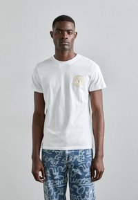 Λευκό βαμβακερό μπλουζάκι με στρογγυλή λαιμόκοψη και κοντά μανίκια, που διαθέτει κυκλικό χρυσό λογότυπο στην αριστερή πλευρά του στήθους. Κλασική εφαρμογή, απλός σχεδιασμός.