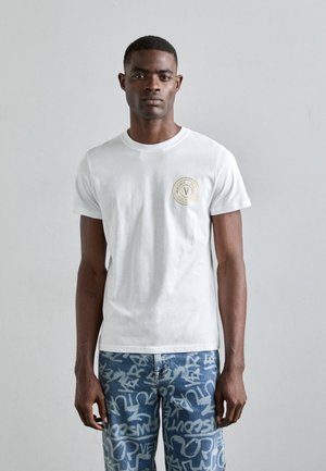 T-shirt bianco in cotone con colletto tondo e maniche corte, con un logo circolare dorato sul lato sinistro del petto. Vestibilità classica, design semplice.