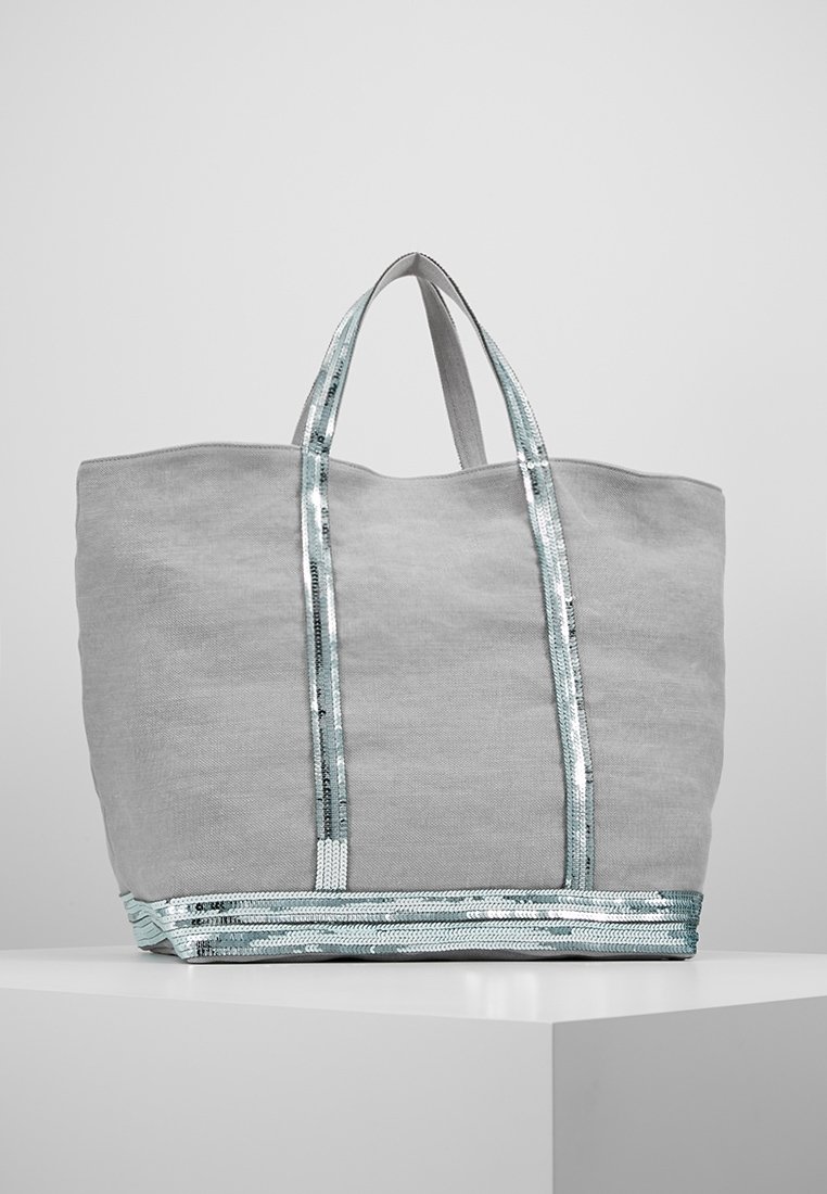 Sac cabas en tissu gris clair avec bordure en sequins argentés brillants sur les anses et le fond, posé sur un piédestal blanc sur fond gris.