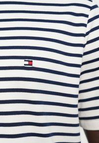 Navy blue and white striped fabric with a small rectangular logo featuring black, white, and red color blocks, centered on the chest.