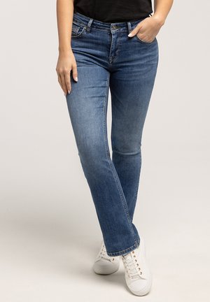 Kobieta ubrana w niebieskie dżinsy straight-leg, sportowe białe trampki, jedna ręka w kieszeni, druga swobodnie przy boku.