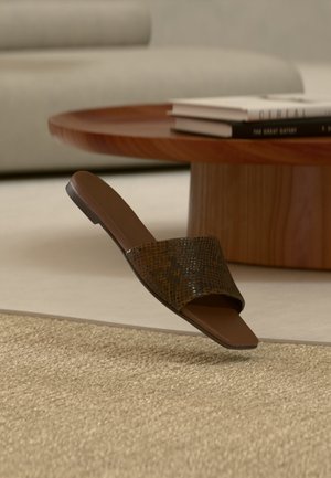 Braune offene Pantolette mit texturierter Riemen auf beigem Teppich, neben einem runden Holzcouchtisch mit gestapelten Büchern.