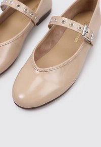 Bailarinas de charol beige tipo Mary Jane con punta redondeada y correa ajustable que presenta un hardware circular plateado, plantilla texturizada.