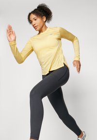 Langermet lys gul treningsskjorte med sideslits, kombinert med tettsittende svarte leggings. Modellen er i en løpeposisjon, som viser aktiv bevegelse.