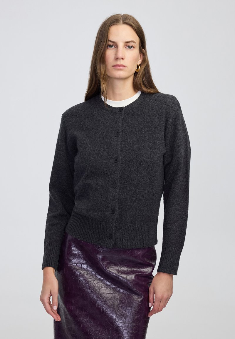 Vrouw met lang bruin haar draagt een donkergrijze geknoopte cardigan, een witte onderhempje, gouden oorringen en een glanzende, gestructureerde donkerpaarse rok, staand.