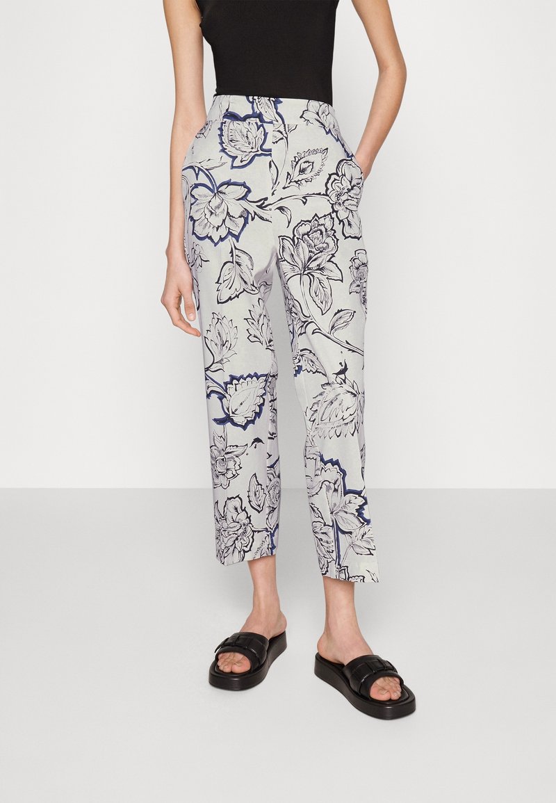 Femme portant un pantalon blanc raccourci avec un grand imprimé floral bleu marine et noir et des sandales noires, debout avec une main dans la poche.
