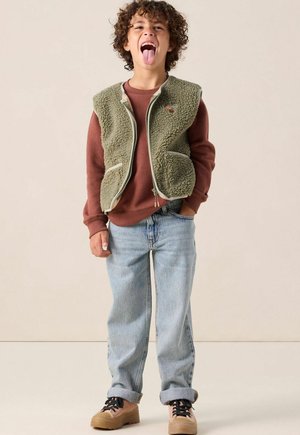 Groene fleece vest met twee voorzakken, gelaagd over een bruine sweatshirt. Gekleed met lichtblauwe jeans en tan schoenen. Kind maakt een speelse gezichtsuitdrukking.